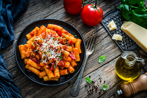 Top view of rigatoni pasta with tomato sauce plate shot on rustic wooden table. Some ingredients like ripe tomatoes, olive oil, basil, peppercorns and Parmesan cheese are all around the plate. XXXL 42Mp studio photo taken with SONY A7rII and Sony FE 90mm f2.8 Macro G OSS lens