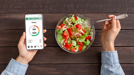 Smart eating and diet planning concept. Woman eating fresh vegetable salad and counting calories on mobile application, top view