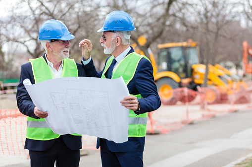 Two senior gray-haired architects or businessmen looking at building plans on a construction site. Engineers meeting and discussing blueprints while visiting work site