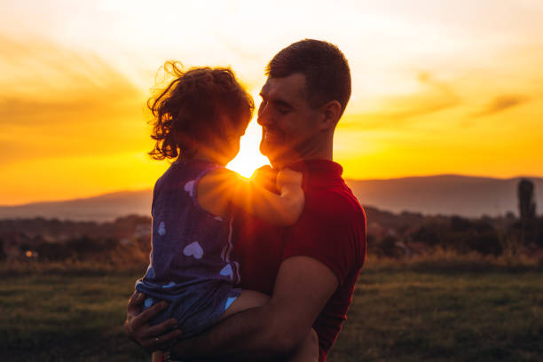 Young caucasian father holding his cute little daughter at his arms at sunset on meadow.