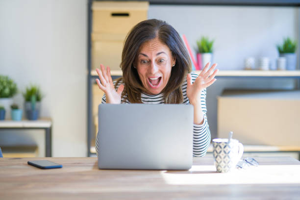 istock Middle age senior woman sitting at the table at home working using computer laptop celebrating crazy and amazed for success with arms raised and open eyes screaming excited. Winner concept 1174112610
