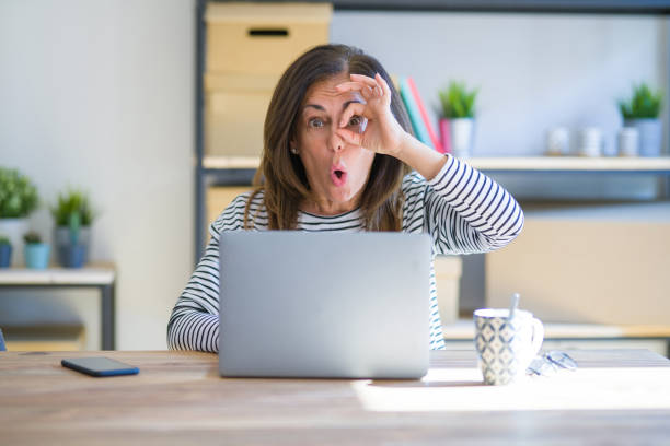 istock Middle age senior woman sitting at the table at home working using computer laptop doing ok gesture shocked with surprised face, eye looking through fingers. Unbelieving expression. 1174112657