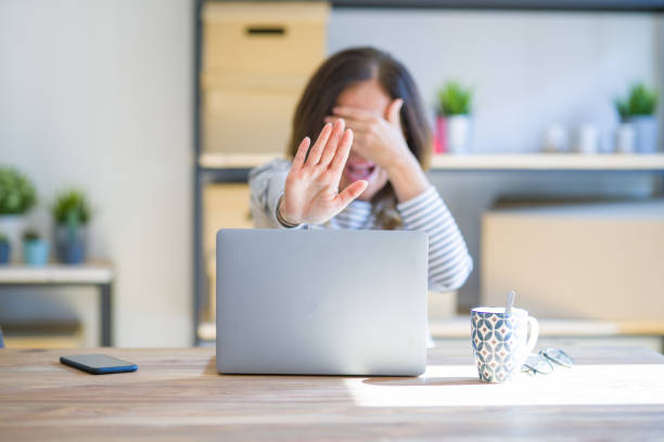 mulher sênior da idade média que senta-se na tabela em casa que trabalha usando os olhos da coberta do portátil do computador com mãos e fazendo o gesto do batente com triste e expressão do medo. conceito embaraçado e negativo. - fear hiding women office - fotografias e filmes do acervo