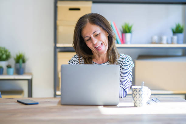 istock Middle age senior woman sitting at the table at home working using computer laptop sticking tongue out happy with funny expression. Emotion concept. 1174112711