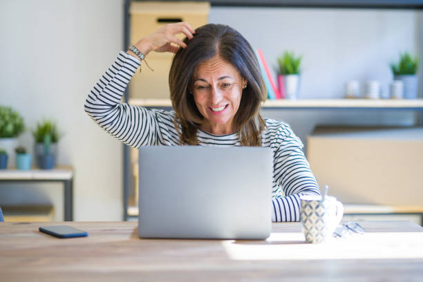 Middle age senior woman sitting at the table at home working using computer laptop confuse and wonder about question. Uncertain with doubt, thinking with hand on head. Pensive concept.