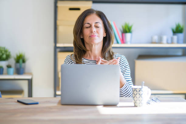 istock Middle age senior woman sitting at the table at home working using computer laptop smiling with hands on chest with closed eyes and grateful gesture on face. Health concept. 1174112805