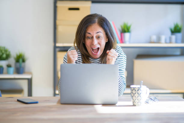 istock Middle age senior woman sitting at the table at home working using computer laptop very happy and excited doing winner gesture with arms raised, smiling and screaming for success. Celebration concept. 1174112838