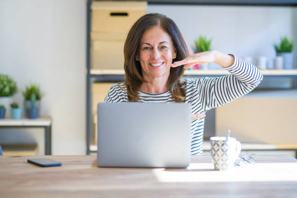 istock Middle age senior woman sitting at the table at home working using computer laptop gesturing with hands showing big and large size sign, measure symbol. Smiling looking at the camera. Measuring concept. 1174112865