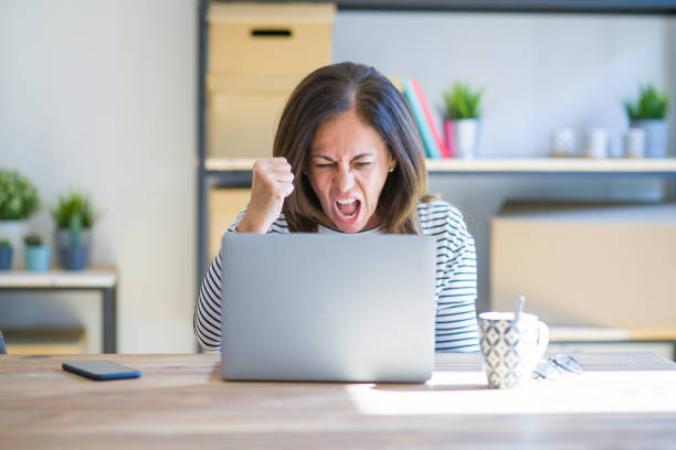 istock Middle age senior woman sitting at the table at home working using computer laptop angry and mad raising fist frustrated and furious while shouting with anger. Rage and aggressive concept. 1174113040