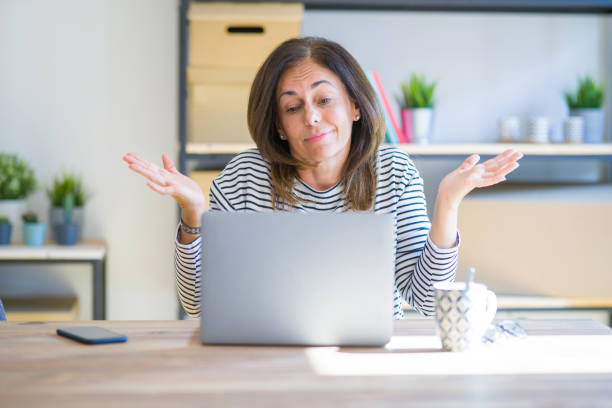 mujer mayor de mediana edad sentada en la mesa en casa trabajando usando computadora portátil despistada y confundida expresión con los brazos y las manos levantadas. concepto de duda. - portrait senior adult confusion women fotografías e imágenes de stock