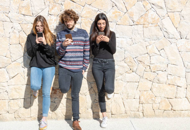 3 people - 1 young man and 2 young women using their smartphones backed by a stone wall - Multiracial and multiethnic group - negative space stock photo