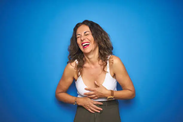 Photo of Middle age senior woman with curly hair standing over blue isolated background smiling and laughing hard out loud because funny crazy joke with hands on body.