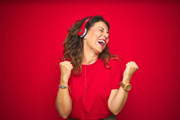 femme aînée d'âge moyen utilisant des écouteurs écoutant la musique au-dessus du fond isolé rouge très heureux et excité faisant le geste gagnant avec les bras levés, souriant et criant pour le succès. concept de célébration. - women female cheerful ecstatic photos et images de collection