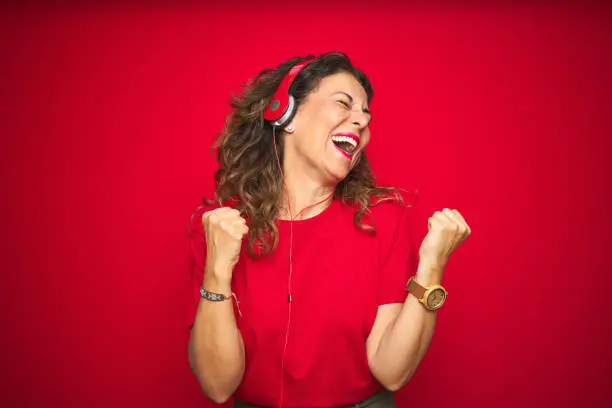 Photo of Middle age senior woman wearing headphones listening to music over red isolated background very happy and excited doing winner gesture with arms raised, smiling and screaming for success. Celebration concept.