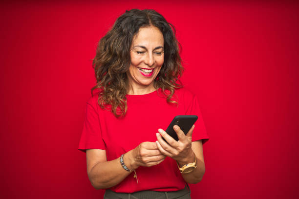 donna anziana di mezza età che usa lo smartphone su sfondo rosso isolato con un viso felice in piedi e sorridente con un sorriso sicuro che mostra i denti - red cell foto e immagini stock