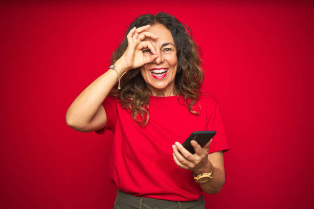 istock Middle age senior woman using smartphone over red isolated background with happy face smiling doing ok sign with hand on eye looking through fingers 1174133366
