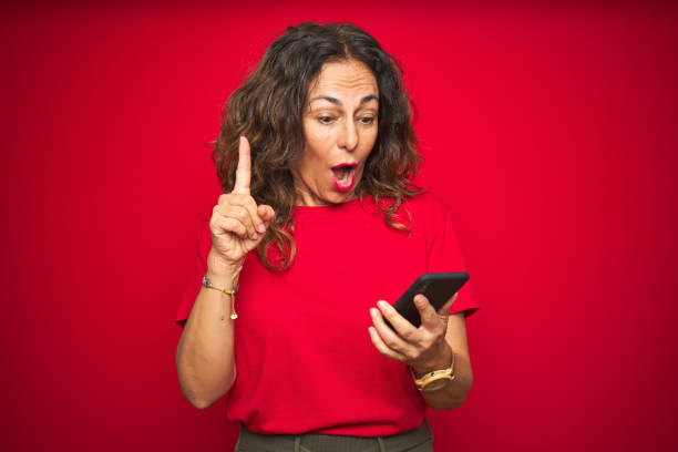 istock Middle age senior woman using smartphone over red isolated background surprised with an idea or question pointing finger with happy face, number one 1174133368