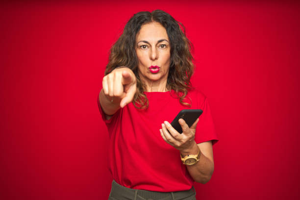 istock Middle age senior woman using smartphone over red isolated background pointing with finger to the camera and to you, hand sign, positive and confident gesture from the front 1174133434