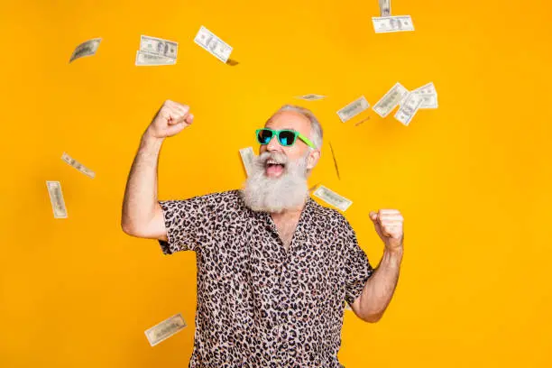 Photo of Portrait of crazy retired funky bearded old man with eyeglasses eyewear raise his fists scream yeah celebrate victory look at money falling wearing leopard shirt isolated over yellow background