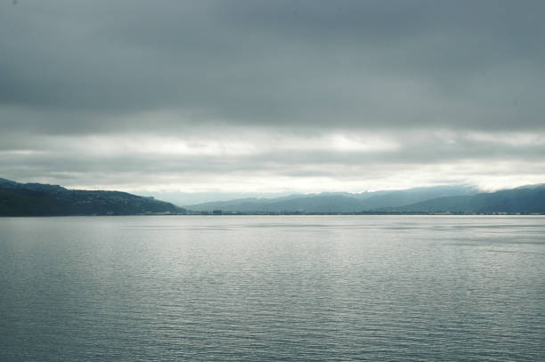 Grey clouds hover over a still lake. The water looks silver. Hills are in the distance, and some sunshine is breaking through the clouds.