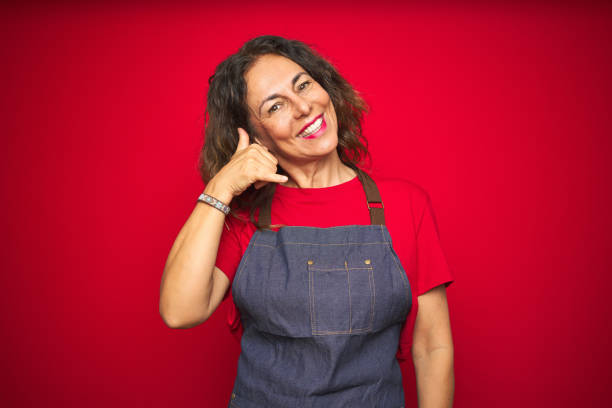 istock Middle age senior woman wearing apron uniform over red isolated background smiling doing phone gesture with hand and fingers like talking on the telephone. Communicating concepts. 1174135856