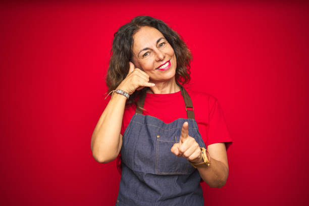 istock Middle age senior woman wearing apron uniform over red isolated background smiling doing talking on the telephone gesture and pointing to you. Call me. 1174136441