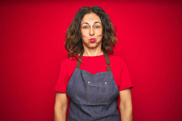 istock Middle age senior woman wearing apron uniform over red isolated background puffing cheeks with funny face. Mouth inflated with air, crazy expression. 1174136529