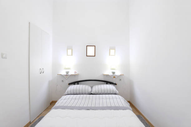 A modern, minimalist bedroom corner featuring a white nightstand with a classic design, a white table lamp, a bed with a white headboard and checkered pillow, a light blue.