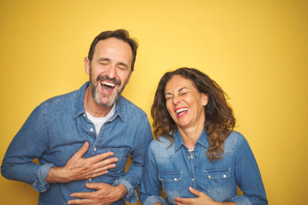 Beautiful middle age couple together standing over isolated yellow background smiling and laughing hard out loud because funny crazy joke with hands on body. Beautiful middle age couple together standing over isolated yellow background smiling and laughing hard out loud because funny crazy joke with hands on body. people laughing hard stock pictures, royalty-free photos & images