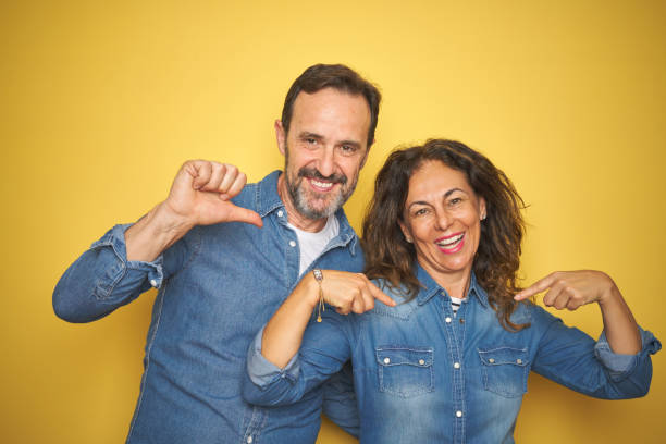 istock Beautiful middle age couple together standing over isolated yellow background looking confident with smile on face, pointing oneself with fingers proud and happy. 1174148416