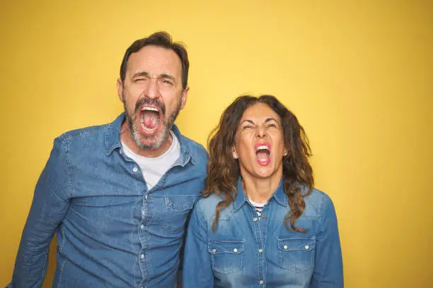 Photo of Beautiful middle age couple together standing over isolated yellow background angry and mad screaming frustrated and furious, shouting with anger. Rage and aggressive concept.