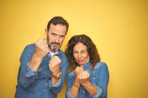 Photo of Beautiful middle age couple together standing over isolated yellow background Ready to fight with fist defense gesture, angry and upset face, afraid of problem