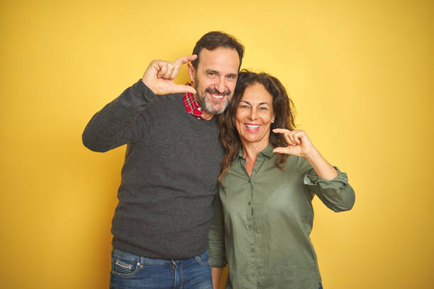 Beautiful middle age couple over isolated yellow background smiling and confident gesturing with hand doing small size sign with fingers looking and the camera. Measure concept. Beautiful middle age couple over isolated yellow background smiling and confident gesturing with hand doing small size sign with fingers looking and the camera. Measure concept. couple isolated wife husband stock pictures, royalty-free photos & images