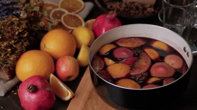 Female hand putting a saucepan of hot mulled wine on cutting board. Fresh lemons, oranges, pomegranate and dried herbs in background. Steam from wine soar over the saucepan