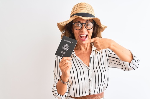 Senior tourist woman wearing hat glasses holding USA passport over isolated white background very happy pointing with hand and finger