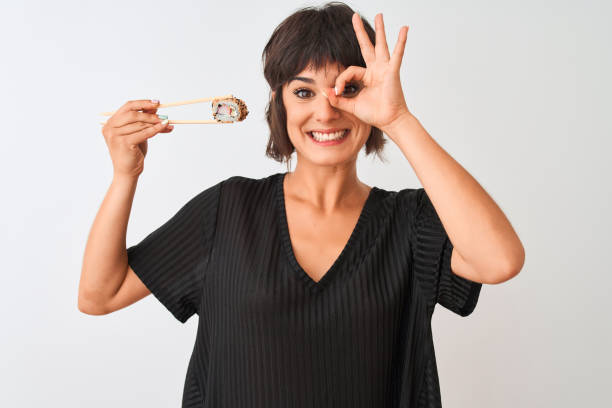 mulher bonita nova que come o sushi usando os chopsticks que estão sobre o fundo branco isolado com sorriso feliz da face que faz o sinal aprovado com mão no olho que olha através dos dedos - chopsticks human hand women isolated - fotografias e filmes do acervo