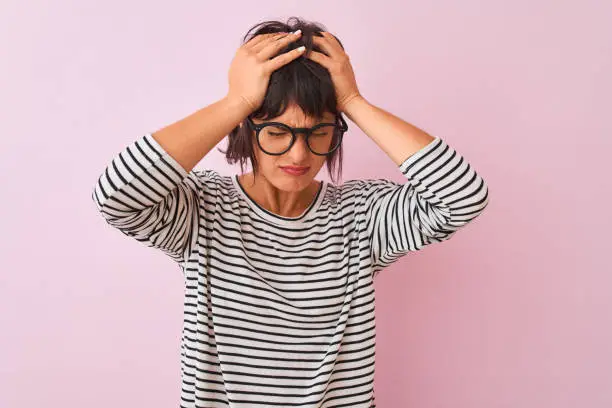 Photo of Young beautiful woman wearing striped t-shirt and glasses over isolated pink background suffering from headache desperate and stressed because pain and migraine. Hands on head.