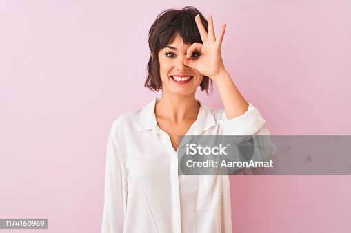 istock Young beautiful woman wearing white shirt standing over isolated pink background doing ok gesture with hand smiling, eye looking through fingers with happy face. 1174160969