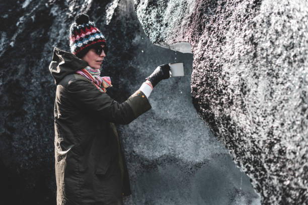 Girl in a khaki jacket surrounded by ice and black sand from the glacier. She hold white metal mug in hands.