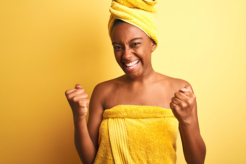 African american woman wearing towel after shower standing over isolated yellow background very happy and excited doing winner gesture with arms raised, smiling and screaming for success. Celebration concept.