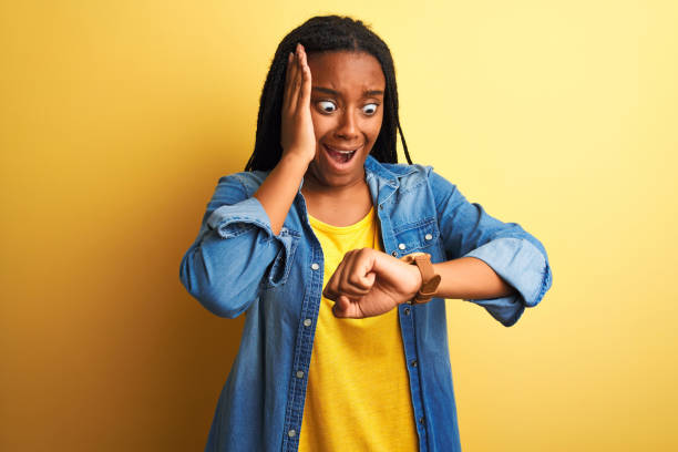 giovane donna afroamericana che indossa una camicia di denim in piedi su uno sfondo giallo isolato guardando il tempo dell'orologio preoccupato, paura di arrivare in ritardo - checking the time women impatient wristwatch foto e immagini stock