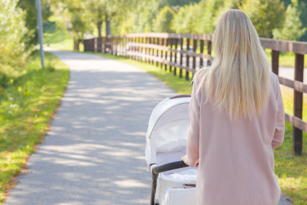 Young blonde mother pushing white baby stroller. Walking at park. Peaceful atmosphere. Spending time with infant in beautiful, sunny summer day. Breathing fresh air. Back view.