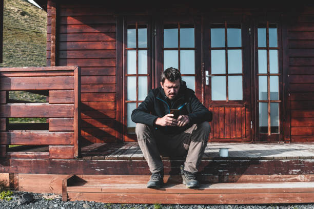 Bearded man in casual attire relaxing on a wooden bench at a scenic forest campsite, holding a metal mug, surrounded by sunlight and peaceful natural scenery.