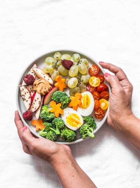 istock Female hands holding a large snack plate. Boiled egg, broccoli, carrots, apples with peanut butter, grapes - delicious diet breakfast, snack on a light background, top view 1174207818