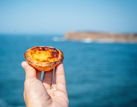 pastel de nata in hand on a background of blue ocean, traditional portuguese food and desserts, travel to europe