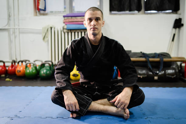 junge bjj brasilianischejiu jitsu athlet jujitsu kämpfer sitzt auf dem tatami-matten boden auf dem training tragen schwarze kimono gi und gürtel - ju jitsu stock-fotos und bilder