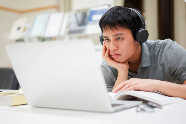 close up young asian man hand on chin with feeling bored while listening online course from webinar for midterm or final test at library room , education lifestyle people concept