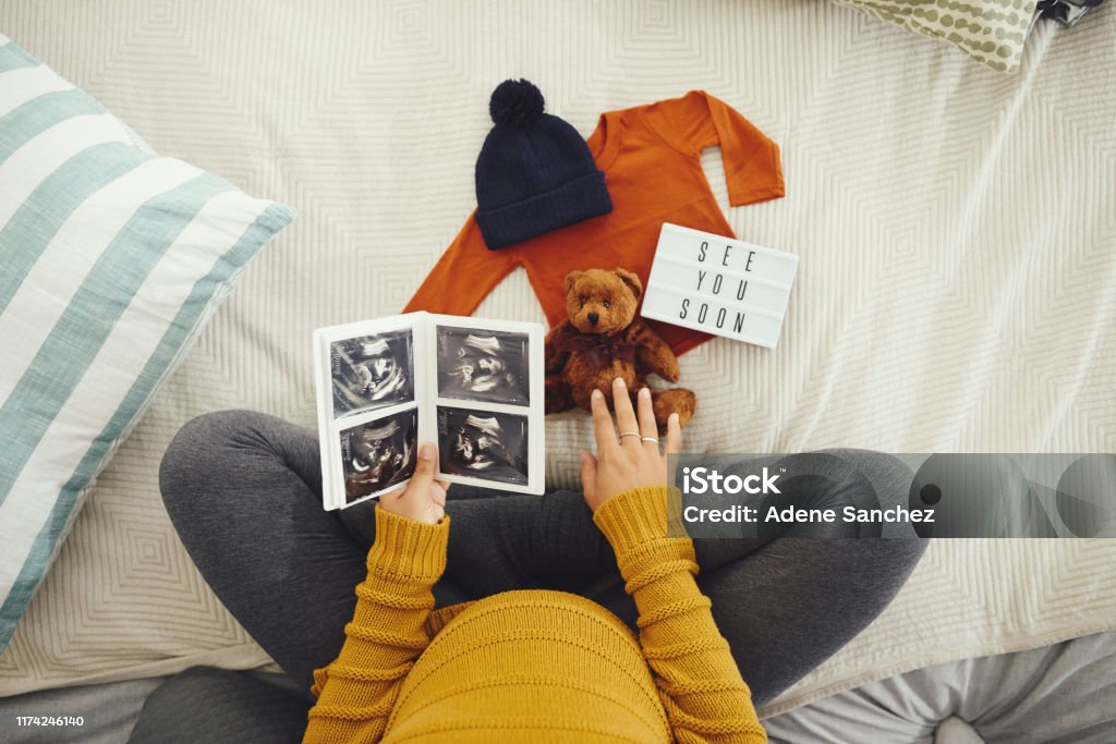 These are the most precious moments in life High angle shot of an unrecognizable pregnant woman looking at a sonogram and baby clothing in her bedroom at home Pregnant Stock Photo