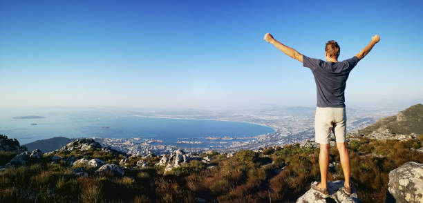 Panoramic rear view of tall man with open arms overlooking Cape Town from the top of table mountain.