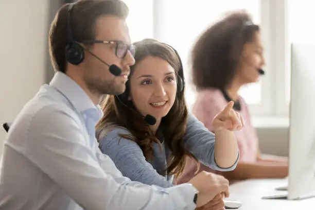 Photo of Female worker in headset help colleague with pc